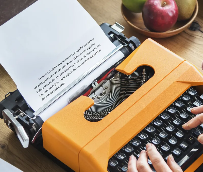 Hands typing a page on an orange typewriter at a wooden desk